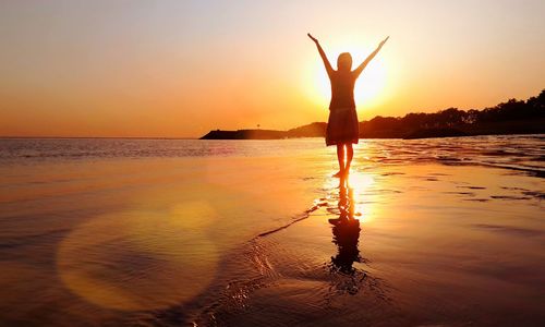 Silhouette woman with arms raised standing at beach during sunset