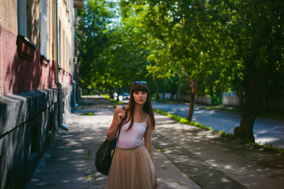 Portrait of young woman standing on sidewalk against trees