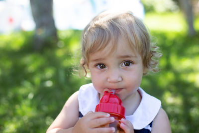 Cute toddler girl with big eyes drinking water from baby bottle outdoors in sunny summer day