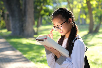 Girl reading book while standing against trees