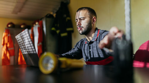 Concentrated young bearded ethnic guy wearing shirt sitting at table with laptop and transmitter looking at screen with yellow flashlight in foreground