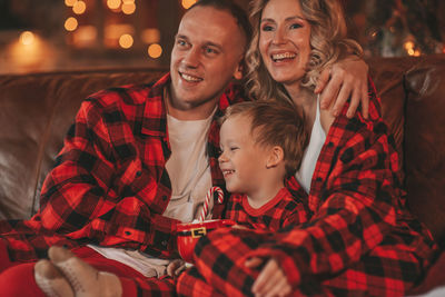 Smile active parents with small son in red checkered sleepwears waiting santa indoor.