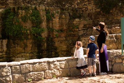 Rear view of woman pointing while standing by boy and girl against stone wall