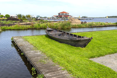 An old wooden boat standing on the shore on the grass, in the background a restaurant and a lake.