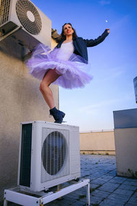 A ballerina in tutu, jacket and boots climbed on the air conditioner and poses against the sky