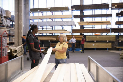 Saleswoman smiling while explaining female customer holding plank in trailer at hardware store