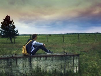 Side view of man sitting on retaining wall at grassy field against cloudy sky