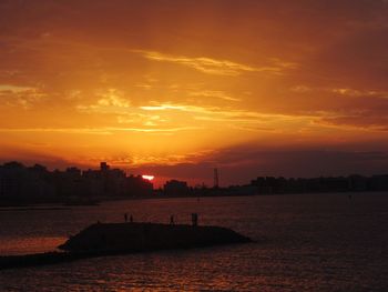 Silhouette buildings by sea against sky during sunset