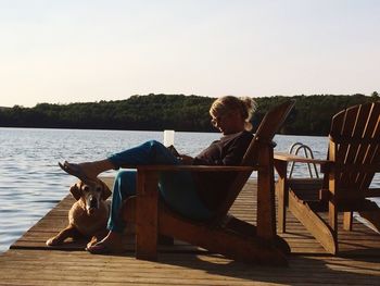 Side view woman with dog sitting on chair at lake against sky