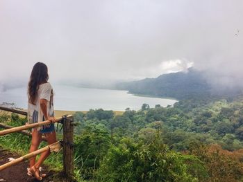 Woman standing by fence while looking at lake and forest during foggy weather