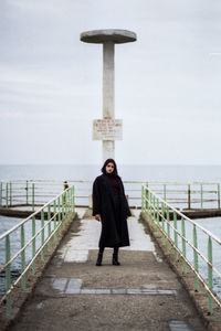 Portrait of young woman standing on pier over sea against cloudy sky