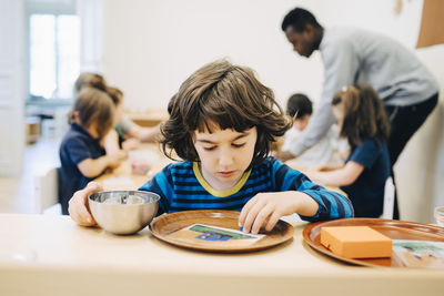 Boy looking at plate on table against friends and teacher in classroom