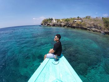 Young man sitting by sea against blue sky