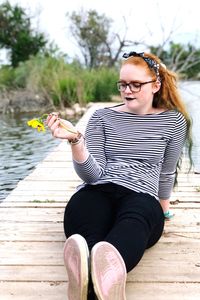 Full length of young woman holding yellow flower while sitting on pier