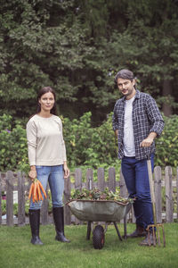 Full length portrait of mid adult couple with bunch of carrots standing by wheelbarrow at organic farm