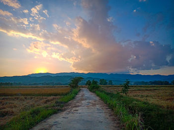 Dirt road amidst field against sky during sunset