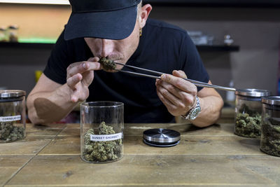 Unrecognizable adult male in cap with tweezers smelling dried hemp floral bud above table with jars in workspace