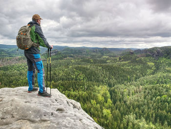 The body of man with a backpack and running shoes stands on top of a rock against the sky