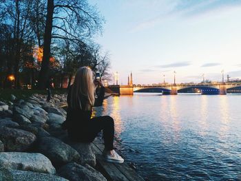 Full length of woman photographing illuminated bridge using smart phone while sitting at riverbank during sunset