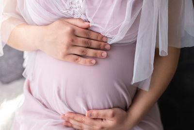 The belly of a pregnant woman in a pink dress holds her hands