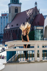 Playful young woman tossing hair on balcony against buildings in city