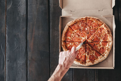 Male hand cuts pepperoni pizza with a round knife on a dark wooden background. 