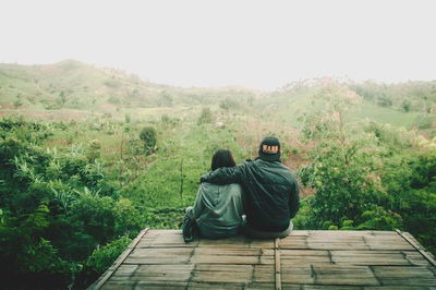 Rear view of couple sitting on wooden structure by trees against sky
