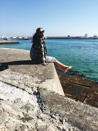 Woman sitting on retaining wall by sea against clear sky