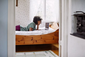 Side view of young woman using laptop while lying on bed at home seen through doorway