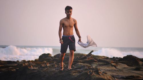 Shirtless mid adult man with tank top walking at beach against clear sky