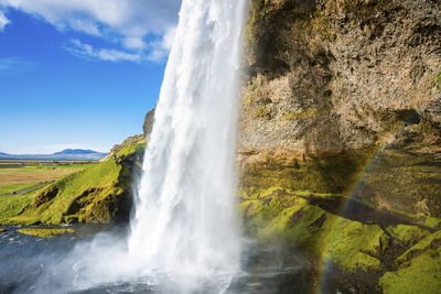 View of seljalandsfoss with rainbow flowing from mountain against cloudy sky