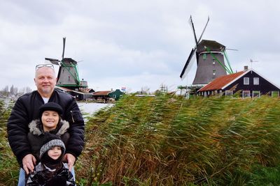 Portrait of father standing with children with traditional windmills in background against cloudy sky