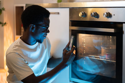 Smiling black guy sitting near oven in kitchen capturing photo of homemade cake baking in oven