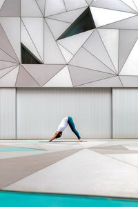 Side view of adult male in sportswear doing downward facing dog exercise while doing yoga against geometric wall in spacious room
