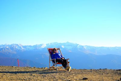 Girl resting on deck chair by mountains against clear blue sky