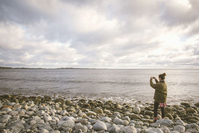 Woman photographing sea against cloudy sky while standing on shore at beach