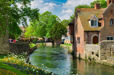 Houses by river amidst trees and buildings against sky