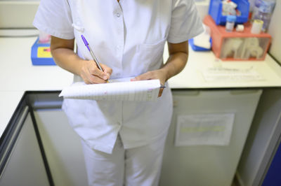 Woman in medical uniform writing on clipboard while standing in office during shift in children hospital