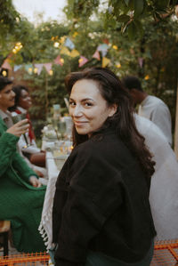 Portrait of smiling woman with long hair looking over shoulder while sitting with friends at dining table in back yard