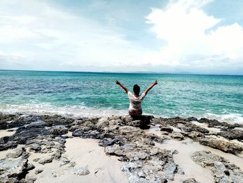 Rear view of woman gesturing peace sign while sitting on shore against sky