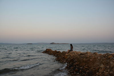 Woman sitting on rock by sea against sky during sunset