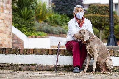 Full length of senior wearing mask with dog sitting outdoors