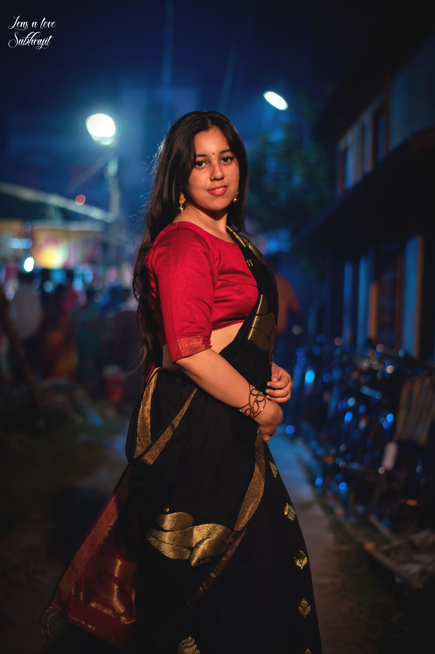 PORTRAIT OF YOUNG WOMAN STANDING ON STREET AT NIGHT