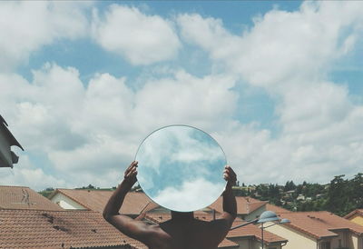 Rear view of shirtless man holding mirror in town against sky