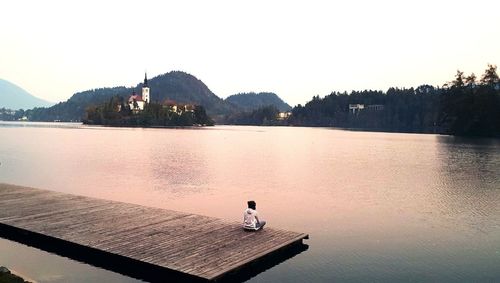 Rear view of man sitting on pier over lake against sky