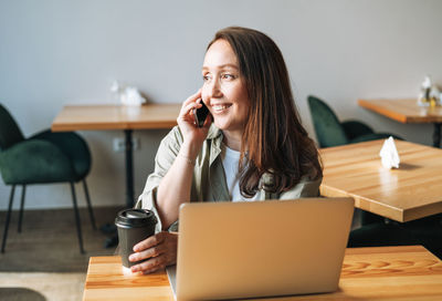 Adult business woman forty years with long hair  shirt working on laptop using mobile phone in cafe