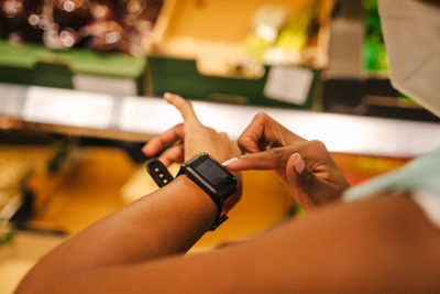 Modern african american female buyer in protective mask for coronavirus prevention using smart watch while standing near stall with grocery products in supermarket