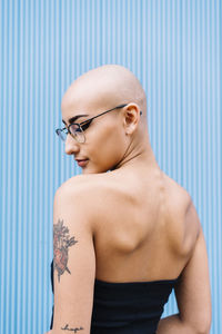 Woman with shaved head wearing eyeglasses in front of blue wall