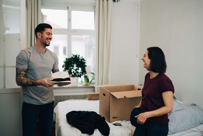 Happy man with hat talking to woman folding clothes by bed at home