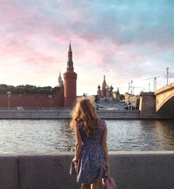 Rear view of mid adult woman standing by river against cloudy sky in city during sunset